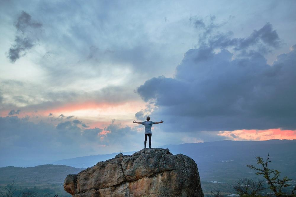 Joshua Earle's photo of the back of a man with arms out wide on top of a boulder that is overlooking a cloudy landscape with the sun setting.