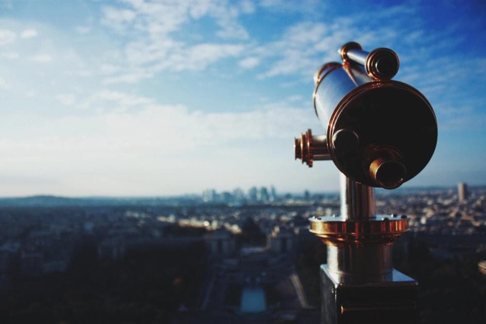 A telescope on top of a high viewpoint is in focus, overlooking the blurry skyline in the distance under a bright blue sky with patchy clouds.